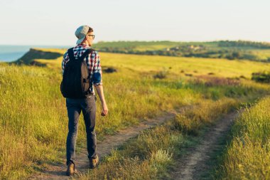 Gülümseyen şık genç adam, gözlük takıyor ve şapka takıyor, yazın sırt çantasıyla seyahat ediyor, yürüyüş yapıyor ve manzarayı gözlemliyor, güzel doğayla iç içe geçmiş hayat tarzı fotoğrafı çekiyor.