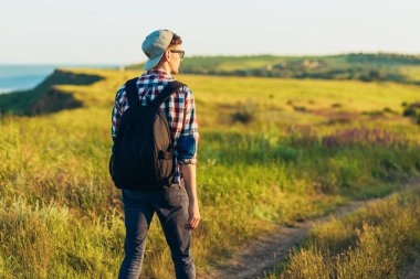 Aktif turist, evrak çantası olan bir adam, kepli bir hippi yeşil bir tepenin üzerinde yürüyor, sağlıklı bir yaşam tarzı, doğa, dinlenme