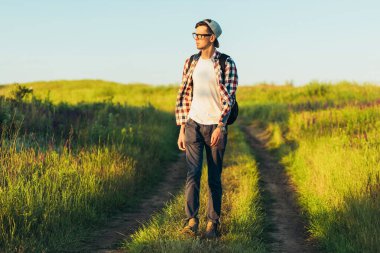 Aktif turist, evrak çantası olan bir adam, kepli bir hippi yeşil bir tepenin üzerinde yürüyor, sağlıklı bir yaşam tarzı, doğa, dinlenme