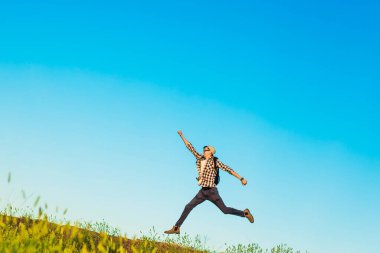 Sırt çantalı mutlu genç bir turist, elleri mavi gökyüzü arka planında zıplayan mutlu bir adam, bir tepenin veya dağın zirvesine tırmanıyor, doğada seyahat, doğa, özgürlük konsepti
