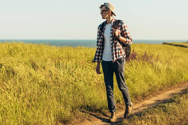 Gülümseyen şık genç adam, gözlük takıyor ve şapka takıyor, yazın sırt çantasıyla seyahat ediyor, yürüyüş yapıyor ve manzarayı gözlemliyor, güzel doğayla iç içe geçmiş hayat tarzı fotoğrafı çekiyor.