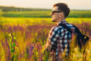 Güneş gözlüklü genç adam kır çiçekleriyle bir tarlada oturuyor gün batımında kırsalda, doğanın, seyahat konseptinin, doğanın tadını çıkarıyor.