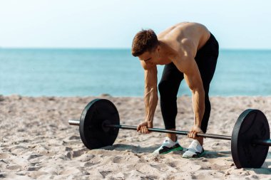 Kas yığını vücut geliştirici. Sahilde büyük dambıllarla egzersiz yapıyor. Güneşli bir yaz gününde, arka planda deniz kıyısında antrenman yapıyor.