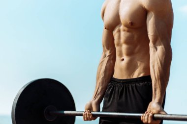 Deniz kenarında kaslı bir adam deniz kenarında antrenman yapıyor, pazılar için halterle egzersiz yapıyor, çıplak gövdeli güçlü bir erkek baskısı, açık hava eğitimi, spor ve spor konsepti.