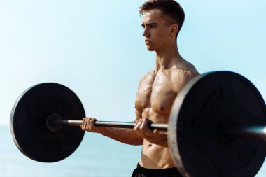 Kas yığını vücut geliştirici. Sahilde büyük dambıllarla egzersiz yapıyor. Güneşli bir yaz gününde, arka planda deniz kıyısında antrenman yapıyor.