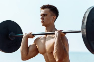Spor giyimli genç adam egzersiz sırasında dambıl kaldırmaya, halterle çömelmeye, deniz kıyısında spor yapmaya odaklandı.