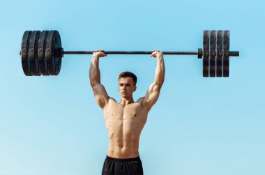 Güzel, şişkin vücutlu kaslı bir adam çıplak bir gövdeyle, halteri kaldırıyor, mavi gökyüzünün arka planında, açık havada egzersiz yapıyor.