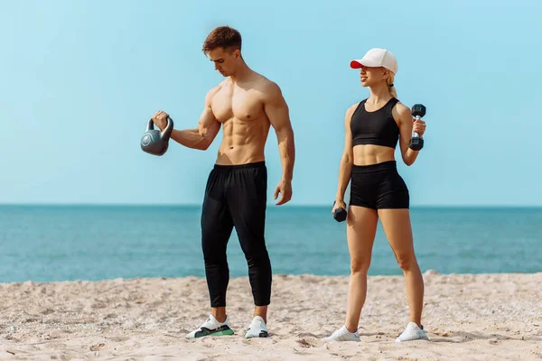 Beautiful young sportive couple exercising with dumbbells, doing sports exercises on the beach, Sports couple training with dumbbells, Muscular man and woman showing muscles