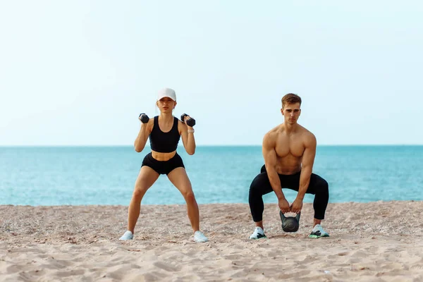 Beautiful young sportive couple exercising with dumbbells, doing sports exercises on the beach, Sports couple training with dumbbells, Muscular man and woman showing muscles