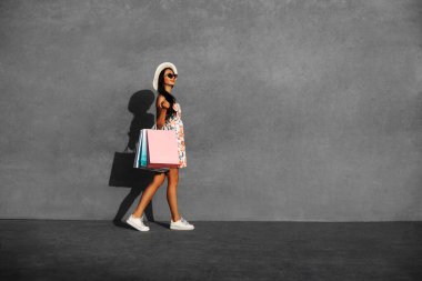 Şık bir kadın, şapka ve güneş gözlüğü takmış, alışveriş çantalarıyla şehir sokaklarında yürüyor, arka planında fotokopi alanı, yaz tarzı, tüketim, alışveriş, yaşam tarzı konsepti olan gri bir şehir duvarına karşı.