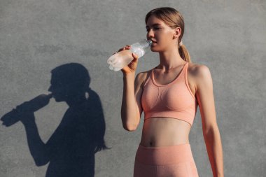Spor giyimli genç bir kadın, dışarıda duvarın yanında duruyor, spor, spor ve motivasyondan sonra bir şişe suyla rahatlatıcı bir kadın.