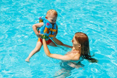 Mutlu bir aile deniz kenarındaki tropikal bir tatil köyünde havuzun mavi sularında oynuyor anne ve küçük bebeği güneşli bir yaz gününde havuzda yüzüyor yaz, dinlenme, tatil, seyahat konsepti.