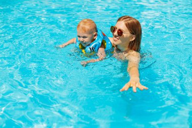 Mutlu aile, anne ve küçük bebek havuzda yüzer, mutlu bebek yüzmeyi öğrenir, havuzda eğlenerek suya dalarlar, anne çocuğuna yüzmeyi öğretir, yaz tatili konseptini öğretir.