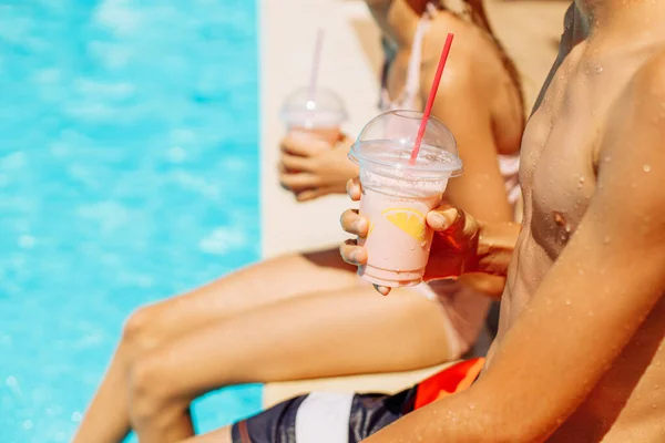 Happy cute kids, boy and girl drinking cool summer cocktails, having fun and relaxing sitting near the pool with clear blue water, happy summer, friendly family weekend concept, outdoor lifestyle