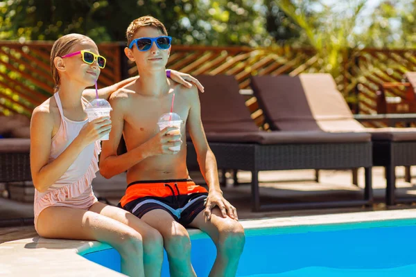 Happy cute kids, boy and girl drinking cool summer cocktails, having fun and relaxing sitting near the pool with clear blue water, happy summer, friendly family weekend concept, outdoor lifestyle