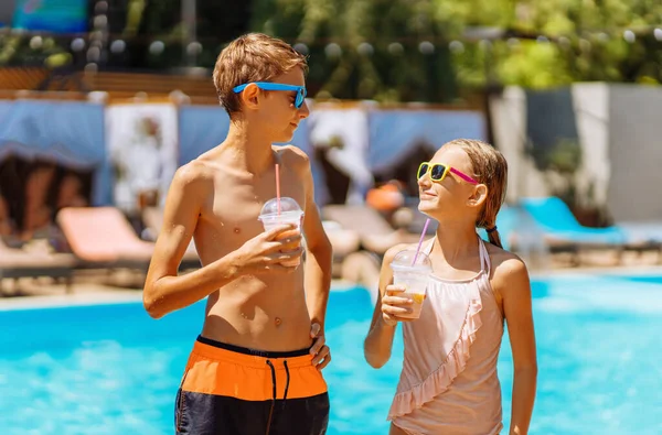 Happy cute kids, boy and girl drinking cool summer cocktails, having fun and relaxing sitting near the pool with clear blue water, happy summer, friendly family weekend concept, outdoor lifestyle