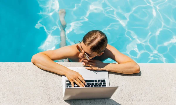 Attractive young woman in sunglasses, uses laptop and work remotely over the pool, Attractive woman smiling, using laptop in the pool, Summer vacation concept, remote work