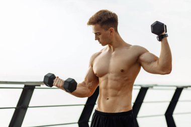 Kas yığını yakışıklı, gövdesi çıplak, halterli güçlü bir adam. İskeledeki köprüde altı kas küpünü gösteriyor.