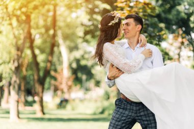 Mutlu düğün çifti, damat mutlu geline hayran, gelini kollarında tutuyor ve onu parka götürüyor, güneşli bir yaz gününde, mutlu bir düğün günü.