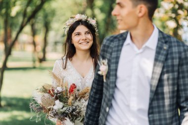 Düğün, mutlu genç damat ve güzel gelinlik içinde bir buketle gelin, gelin ve damat el ele tutuşup yeşil bir tarlada yürüyorlar, düğün günü, boho tarzında düğün töreni.