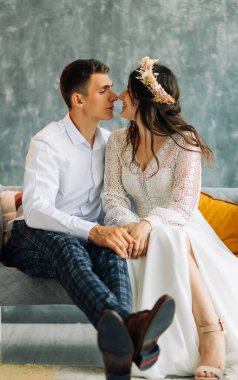 Gri takım elbiseli genç bir damat ve beyaz dantelli bir gelinlik içinde güzel bir gelin, düğün çifti gri bir arka planda koltukta oturuyor, birbirine aşık bir çiftin düğün portresi.