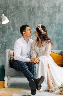 Gri takım elbiseli genç bir damat ve beyaz dantelli bir gelinlik içinde güzel bir gelin, düğün çifti gri bir arka planda koltukta oturuyor, birbirine aşık bir çiftin düğün portresi.