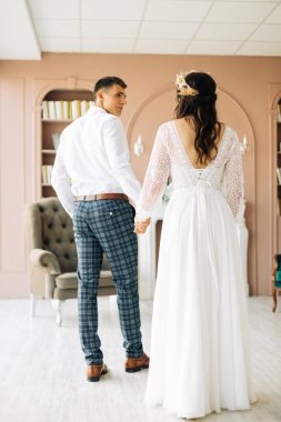 Mutlu genç bir çift, takım elbiseli şık bir damat ve bir buket çiçekli dantelli gelinlik içinde genç bir gelin, birlikte oturur, sarılırız, stüdyoda kanepede, boho tarzı bir düğünle.