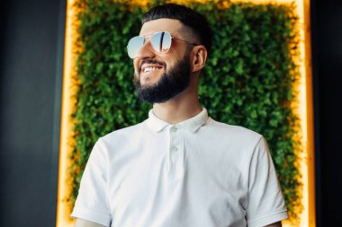 Beyaz gömlek ve güneş gözlüğü takmış, sakallı, mutlu bir gencin portresi, duvarda yeşil yapay çimenler var.