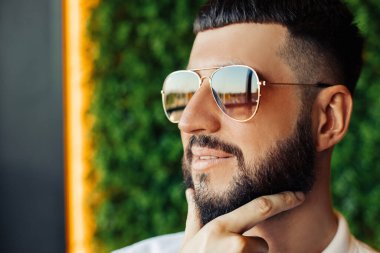 Sakallı, güneş gözlüklü ve beyaz gömlekli mutlu bir adam. Kameraya bakarken gülümseyen genç bir adamın portresi.