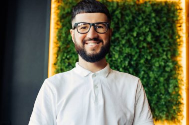 Beyaz tişörtlü yakışıklı, kendine güvenen genç bir adam kameraya bakıyor ve gülümsüyor, bir kafede oturuyor, dinleniyor ve kafede iyi vakit geçiriyor.