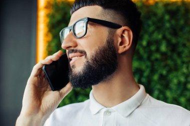 Beyaz gömlekli, gözlüklü, cep telefonuyla konuşan ve kafede otururken gülümseyen genç adam kahve molasında bir kafede oturuyor.