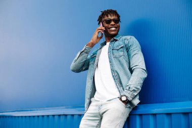 Arayan Afrikalı bir adam mavi arka planda, 5G internet konsepti, telefondaki yüksek hızlı internet, elinde telefonla gülen mavi tişörtlü siyah bir adam.