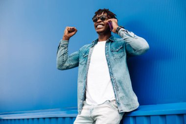 Başarılı Amerikalı siyahi adam telefonla arıyor, zafere giden yolda başarı ve sevinç belirtileri gösteriyor, mavi arka planda