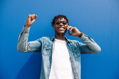 Başarılı Amerikalı siyahi adam telefonla arıyor, zafere giden yolda başarı ve sevinç belirtileri gösteriyor, mavi arka planda