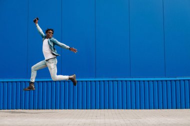 Afrikalı bir adam elinde telefonla şehirde mavi bir binanın arka planına atlıyor. Başarı kavramı.