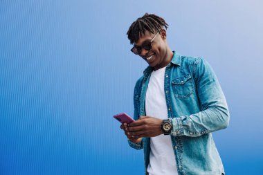 Güneş gözlüklü Afrikalı bir adam mavi arka planda SMS yazıyor, 5G internet konsepti, telefonda yüksek hızlı internet, mavi çuvallı siyah adam, ve kot pantolon.