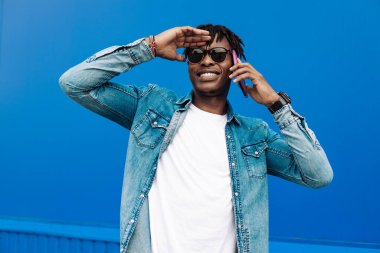 Gözlüklü Afrikalı bir adam mavi arka planda SMS yazıyor, 5G internet konsepti, mesafeye bakıyor, mavi gömlekli bir adam elinde telefonla gülüyor.