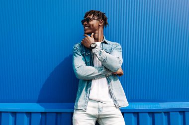 Moda meraklısı, güneş gözlüklü modern elbiseli bir adam mavi bir binanın arka planında poz veriyor.