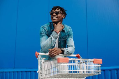 Alışveriş arabasıyla afro alışverişi, alışveriş merkezinin yanında alışveriş yapan adam, mavi bir binanın arka planında genç bir siyahi öğrenci.