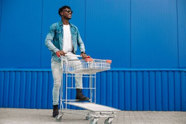 Alışveriş arabasıyla afro alışverişi, alışveriş merkezinin yanında alışveriş yapan adam, mavi bir binanın arka planında genç bir siyahi öğrenci.