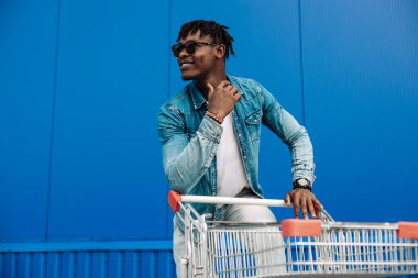 Alışveriş arabasıyla afro alışverişi, alışveriş merkezinin yanında alışveriş yapan adam, mavi bir binanın arka planında genç bir siyahi öğrenci.