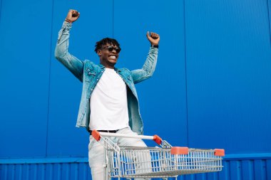 Alışveriş arabasıyla afro alışverişi yapıyor, alışveriş merkezinin yakınında alışveriş yapıyor, seviniyor ve ellerini kaldırıyor, konsept: başarılı satın alma