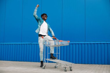 Alışveriş arabasıyla afro alışverişi yapıyor, alışveriş merkezinin yakınında alışveriş yapıyor, seviniyor ve ellerini kaldırıyor, konsept: başarılı satın alma