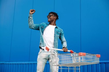 Alışveriş arabasıyla afro alışverişi yapıyor, alışveriş merkezinin yakınında alışveriş yapıyor, seviniyor ve ellerini kaldırıyor, konsept: başarılı satın alma