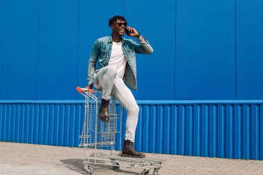 Afrikalı siyahi bir adam alışveriş arabasında oturmuş telefonla konuşuyor, internetten alışveriş yapıyor, mavi bir binanın arka planında bir operatörle konuşuyor.