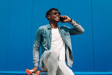 Afrikalı siyahi bir adam alışveriş arabasında oturmuş telefonla konuşuyor, internetten alışveriş yapıyor, mavi bir binanın arka planında bir operatörle konuşuyor.