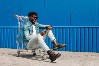 Elinde telefon, elinde alışveriş arabası olan Afro siyahi bir adam mavi bir binanın arka planında internetten alım yapıyor.