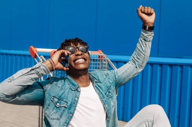 Afrikalı siyahi bir adam alışveriş arabasında oturmuş telefonla konuşuyor, internetten alışveriş yapıyor, mavi bir binanın arka planında bir operatörle konuşuyor.