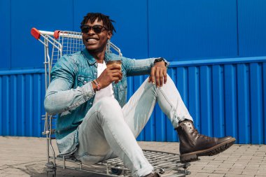 Elinde bir fincan kahve ya da çay tutan Afrikalı siyahi bir adam, alışveriş merkezinin yakınındaki bir alışveriş arabasında, mavi bir binanın arka planında oturuyor.