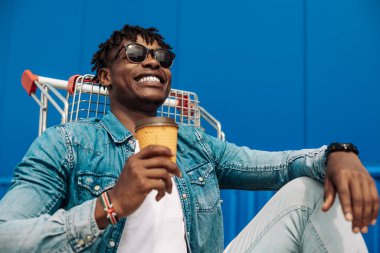 Elinde bir fincan kahve ya da çay tutan Afrikalı siyahi bir adam, alışveriş merkezinin yakınındaki bir alışveriş arabasında, mavi bir binanın arka planında oturuyor.